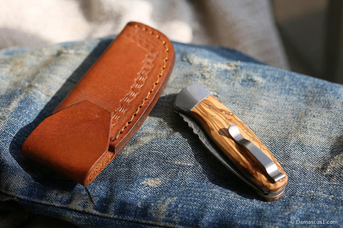 Olive wood Damascus pocket knife closed beside its stitched leather sheath on worn denim, showing how to carry a pocket knife without a clip