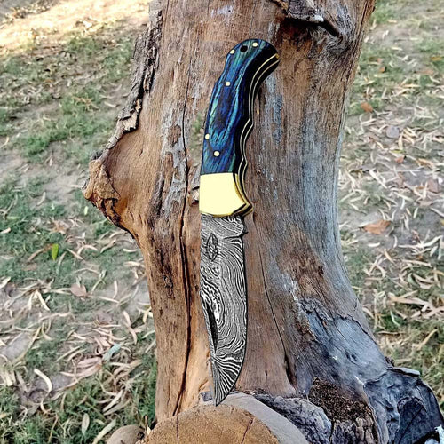 Handmade Damascus steel folding pocket knife with blue wood handle and brass bolster, open against a tree trunk