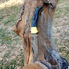 Handmade Damascus steel folding pocket knife with blue wood handle and brass bolster, open against a tree trunk