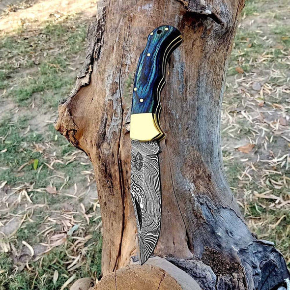 Handmade Damascus steel folding pocket knife with blue wood handle and brass bolster, open against a tree trunk